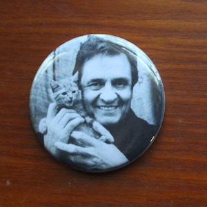 May include: A black and white photo of a man holding a small kitten. The man is smiling and the kitten is looking at the camera.