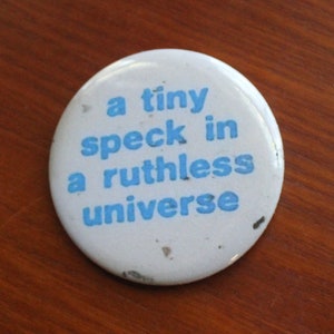 May include: A round white pinback button featuring the blue text 'a tiny speck in a ruthless universe' printed in a sans-serif font. The button is positioned next to a United States quarter coin for scale, showing the button is slightly larger.