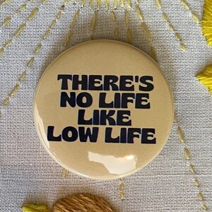 May include: A round, tan pin-back button featuring the dark blue text "THERE'S NO LIFE LIKE LOW LIFE" in a bold, retro font. The button is positioned next to a standard United States quarter coin for scale, showing its relative size.