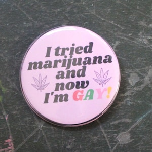 Può includere: Bottone rosa con il testo "Ho provato la marijuana e ora sono GAY!" in testo nero con grafica di foglie di marijuana verdi.