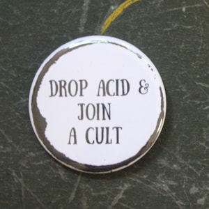 May include: A white button with the text "DROP ACID & JOIN A CULT" in black lettering. The button has a silver edge and a distressed, grunge-style border. A U.S. quarter coin is next to the button for size comparison.