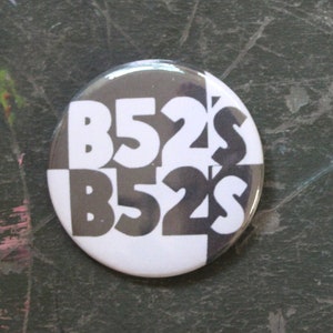 May include: A circular pinback button featuring a black and white graphic design with the text B52'S repeated twice. The button is positioned next to a United States quarter coin for scale, which has a diameter of approximately 0.955 inches.