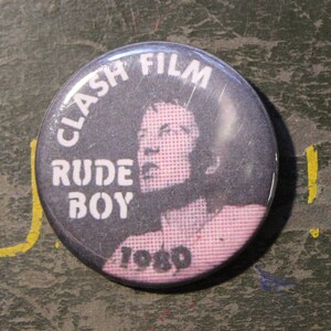 以下が含まれることがあります： 「Clash Film Rude Boy 1980」という文字と、ピンクのハイライトが入った髪の人の白黒写真が描かれた白黒のボタンです。