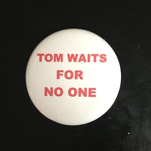 May include: A white button with the text "TOM WAITS FOR NO ONE" in red lettering. A U.S. quarter coin is next to the button. The button is round and appears to be made of metal or plastic.