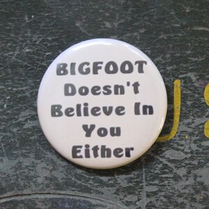May include: A round white pin-back button featuring the black text BIGFOOT Doesn't Believe In You Either, positioned next to a silver United States quarter coin for scale, showing the relative size of the circular accessory.