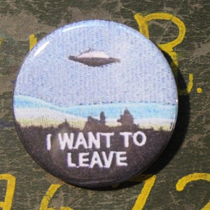 May include: A round button with a blue and white image of a UFO flying over a landscape. The text "I WANT TO LEAVE" is printed in white on a black background.