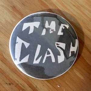 Peut inclure: Un bouton rond noir et blanc avec le texte "THE CLASH" en lettres blanches.