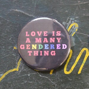 May include: A black button with the text "LOVE IS A MANY GENDERED THING" in rainbow colors. The button is on a green surface.