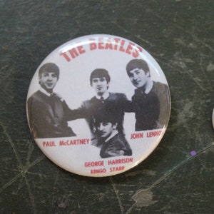 Puede incluir: Un botón blanco con una foto en blanco y negro de los Beatles. El botón tiene texto rojo que dice "The Beatles" y los nombres de los miembros de la banda: "Paul McCartney", "George Harrison", "Ringo Starr" y "John Lennon".