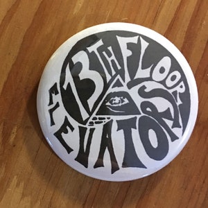 May include: A white button with black text reading "13th Floor Elevators" in a stylized font. The button features an eye within a triangle. A US quarter is next to the button.