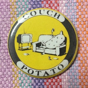 Könnte beinhalten: Ein gelber Knopf mit schwarzem Rand und dem Text "Couch Potato" in Schwarz. Der Knopf zeigt ein Cartoon-Bild einer Kartoffel, die auf einem Sofa sitzt und eine Fernbedienung in der Hand hält.