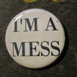 Pode incluir: Um broche redondo branco com o texto preto I'M A MESS impresso no centro. O broche está posicionado ao lado de uma moeda de 25 centavos dos Estados Unidos para escala, que tem um diâmetro de aproximadamente 2,43 cm.
