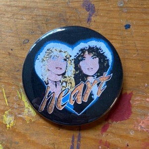 May include: A black button with a blue heart-shaped border featuring two women's portraits and the word "heart" in a stylized font. A U.S. quarter is next to the button for size comparison.