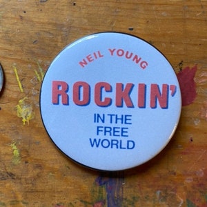 May include: A white button with a blue and red design. The button says "Neil Young Rockin' In The Free World".