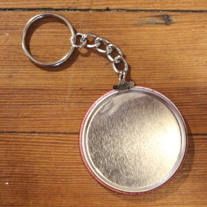 May include: A silver keychain with a round, silver button. The button has a red rim and a smooth, silver surface.