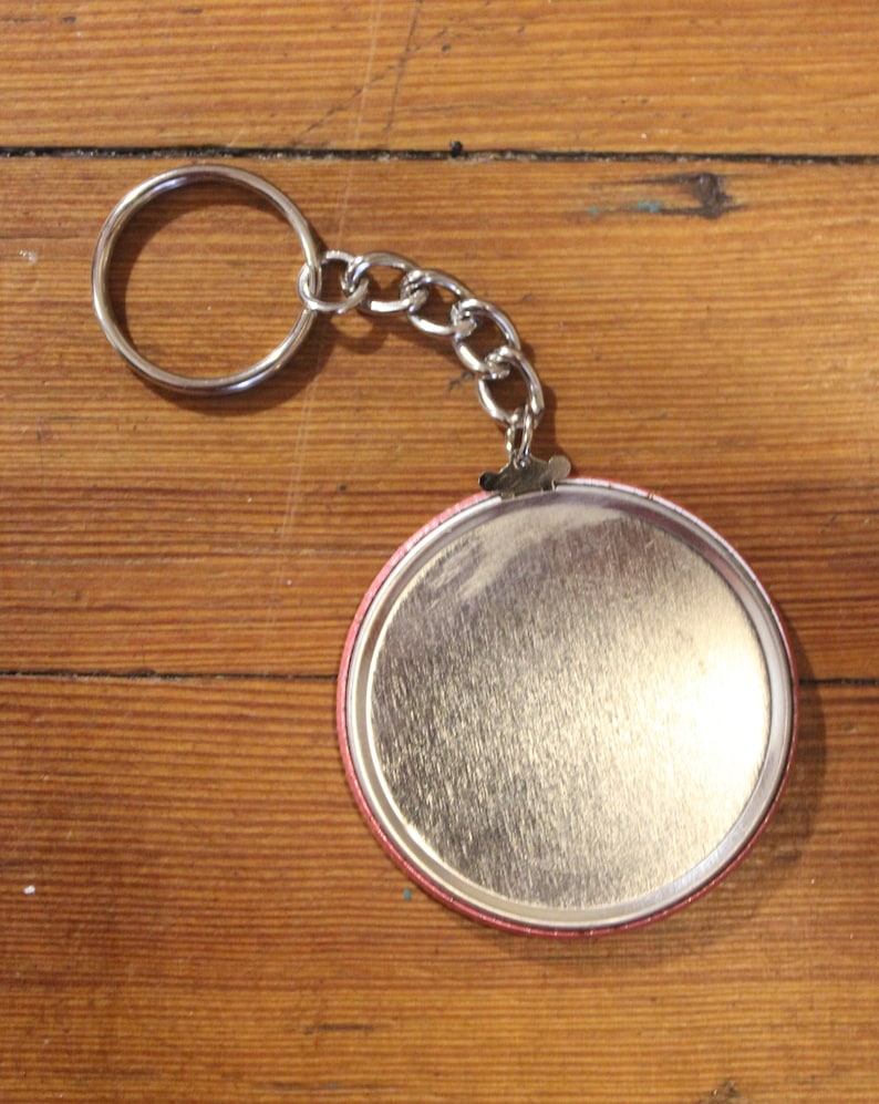 May include: A silver keychain with a round, silver button. The button has a red rim and a smooth, silver surface.