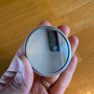 May include: A round, silver-colored compact mirror with a gold trim. The mirror is held in a hand.