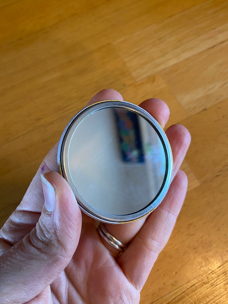 May include: A round, silver-colored compact mirror with a gold trim. The mirror is held in a hand.