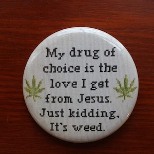 May include: A round white pinback button featuring black cross-stitch style text that reads: My drug of choice is the love I get from Jesus. Just kidding. It's weed. Two small green cannabis leaf illustrations are positioned on the left and right sides.