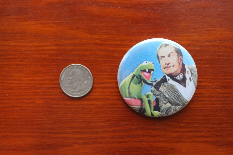 May include: A round button featuring a green Muppet character with a toothy grin, sitting next to a man with a white shirt and a patterned jacket. The background is a light blue sky.