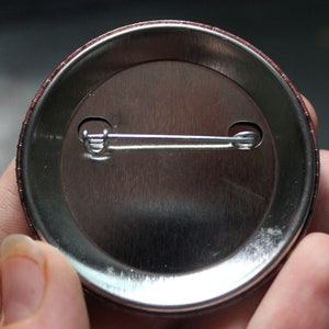 May include: The back of a round button with a metal pin and a brown border.