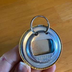 May include: A gold-colored bottle opener keychain with a silver metal ring. The opener has the words "Bottle Opener" printed on it.