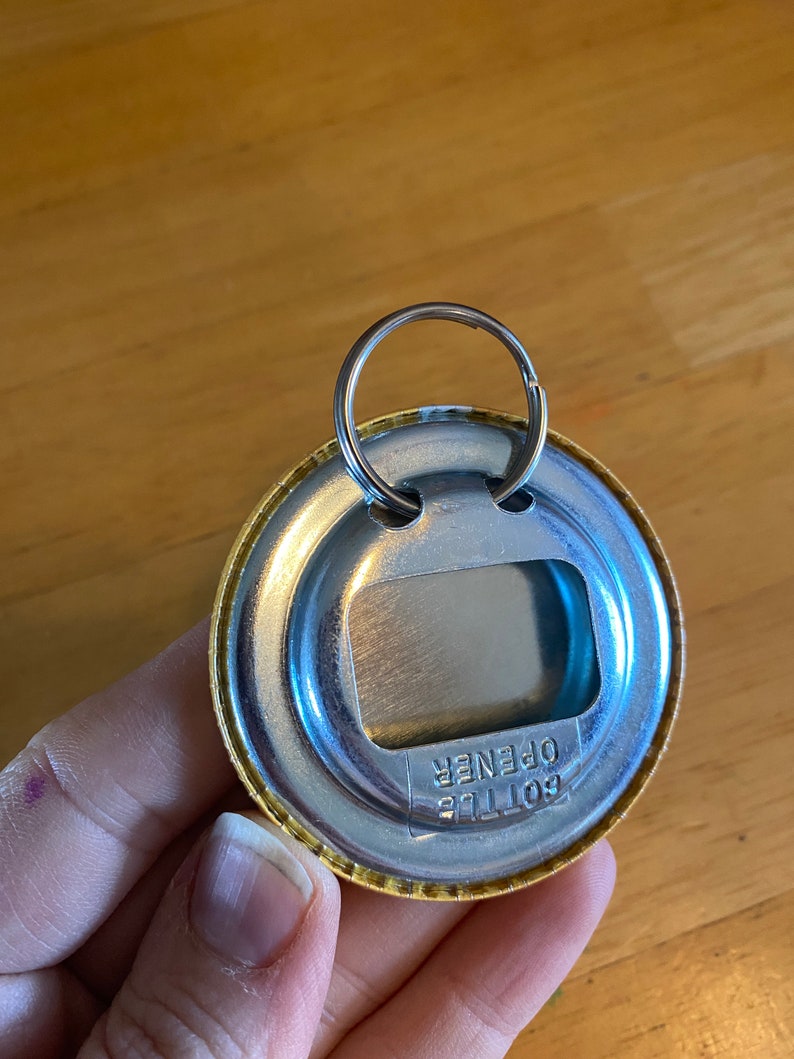May include: A gold-colored bottle opener keychain with a silver metal ring. The opener has the words "Bottle Opener" printed on it.