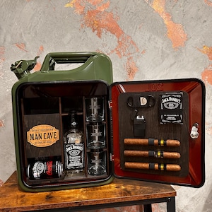 May include: A green metal jerry can converted into a bar cabinet. Inside, there's a bottle of Jack Daniel's whiskey, shot glasses, cigars, a lighter, and a can of Coca-Cola. The words "MAN CAVE" are visible on the wooden interior.