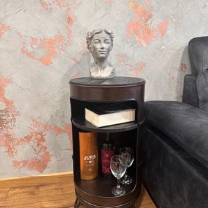 May include: A dark brown metal side table with two shelves. The top shelf holds a small statue of a woman's head. The lower shelves display a bottle of liquor, a bottle of wine, two wine glasses, and a book. The table has a round shape.
