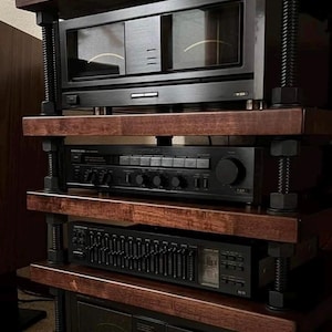 May include: A multi-tiered audio equipment stand with dark wood shelves and black components. The stand is supported by threaded rods and large nuts. The components include a receiver, amplifier, and equalizer, all in black.