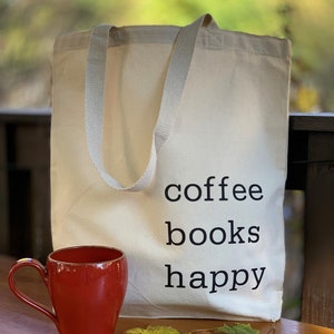 May include: A white canvas tote bag with black text that reads "coffee books happy". A red mug is sitting on a wooden table next to the bag.