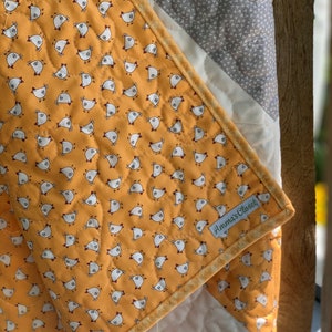 May include: A yellow and white quilt with a pattern of small chickens. The quilt has a gray and white border and a label that says "Anna's Closet".