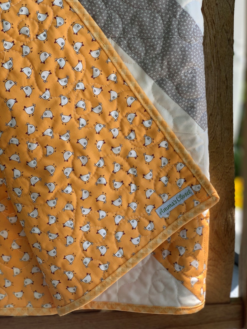 May include: A yellow and white quilt with a pattern of small chickens. The quilt has a gray and white border and a label that says "Anna's Closet".