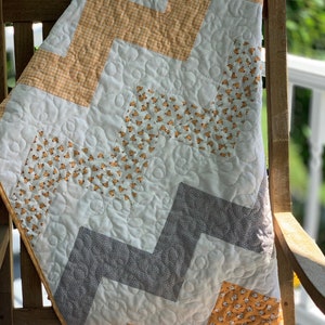 May include: A white, gray, and yellow chevron patterned quilt with a quilted design. The quilt is folded and draped over a wooden chair.