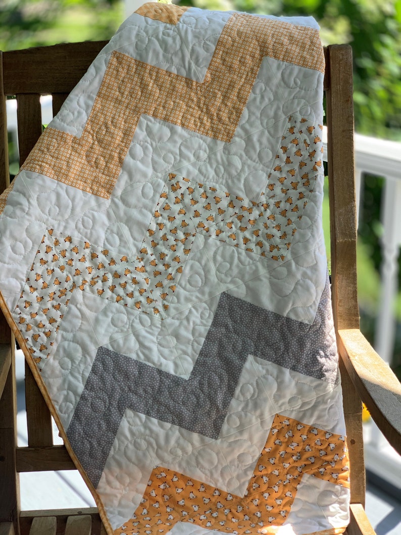 May include: A white, gray, and yellow chevron patterned quilt with a quilted design. The quilt is folded and draped over a wooden chair.