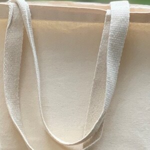 May include: A plain white canvas tote bag with two long, cream-colored straps.