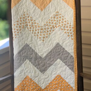 May include: A chevron patterned baby quilt with a white background and alternating rows of gray and yellow fabric. The yellow fabric has a small white chick pattern.