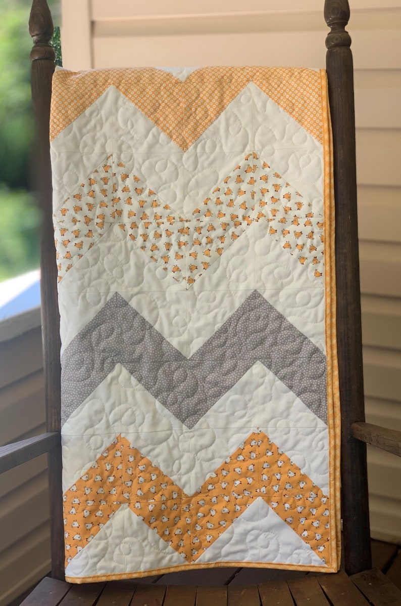 May include: A chevron patterned baby quilt with a white background and alternating rows of gray and yellow fabric. The yellow fabric has a small white chick pattern.
