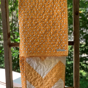 May include: A yellow and white quilt with a chevron pattern and a small chick print on the back. The quilt is hanging on a wooden ladder.