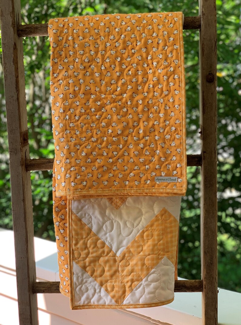 May include: A yellow and white quilt with a chevron pattern and a small chick print on the back. The quilt is hanging on a wooden ladder.