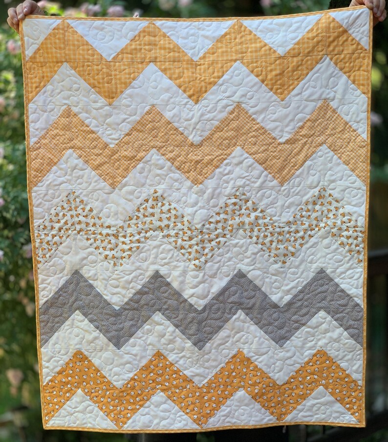 May include: A yellow, gray, and white chevron baby quilt with a small-scale floral print. The quilt is quilted with a swirl pattern.