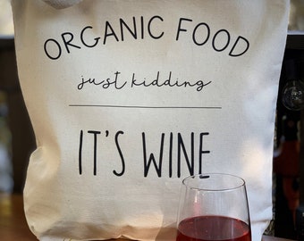 May include: A white canvas tote bag with the text "ORGANIC FOOD just kidding IT'S WINE" printed in black. A glass of red wine sits on a wooden table next to the bag.