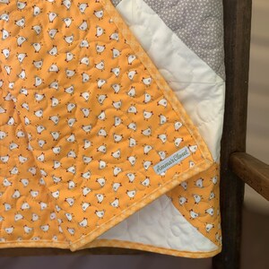 May include: A yellow and white quilt with a pattern of small chickens. The quilt has a gray and white chevron pattern on the back. The quilt has a yellow and white checkered binding and a tag that says "Amma's Closet".