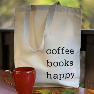 May include: A white canvas tote bag with black text that reads "coffee books happy". A red mug is in the foreground.
