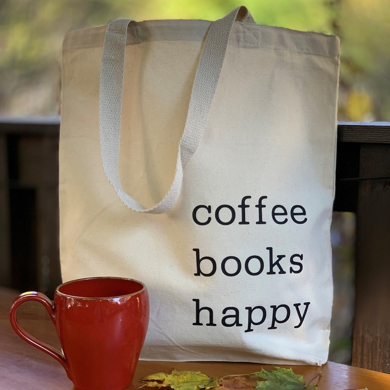 May include: A white canvas tote bag with black text that reads "coffee books happy". A red mug is in the foreground.