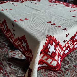 Könnte beinhalten: Cremefarbene Tischdecke mit aufwendigen rot-weißen, gestickten geometrischen Mustern an den Rändern und über die Oberfläche verteilt. Die Tischdecke hat eine gewebte Textur, die ihren rustikalen Charme unterstreicht. Das Design zeigt stilisierte Blumen- und abstrakte Motive.