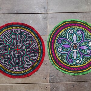 Op de afbeelding: Twee ronde, geborduurde textielstukken met ingewikkelde ontwerpen. Eén heeft een rode rand en geometrische patronen in zwart, wit en groen. De andere heeft een groene rand en een bloemmotief in paars, geel en blauw.