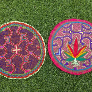 May include: Two round, colorful, embroidered textile pieces with intricate designs. One features a cross-like pattern in pink, green, and yellow on a dark purple background. The other displays a cannabis leaf design in red, yellow, and green.