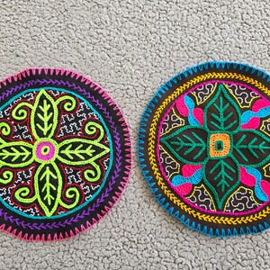 Op de afbeelding: Twee ronde, geborduurde onderzetters met ingewikkelde bloemmotieven. Eén onderzetter heeft een neon groene en roze bloem, de andere heeft een turquoise en roze bloem. Beide staan op een donkerbruine achtergrond met kleurrijke stiksels.