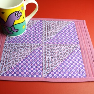 Könnte beinhalten: Ein rosa gesteppter Untersetzer mit einem violetten und weißen Blumenmuster. Der Untersetzer hat einen dekorativen Stich um den Rand und ist perfekt, um Oberflächen vor heißen Tassen zu schützen.